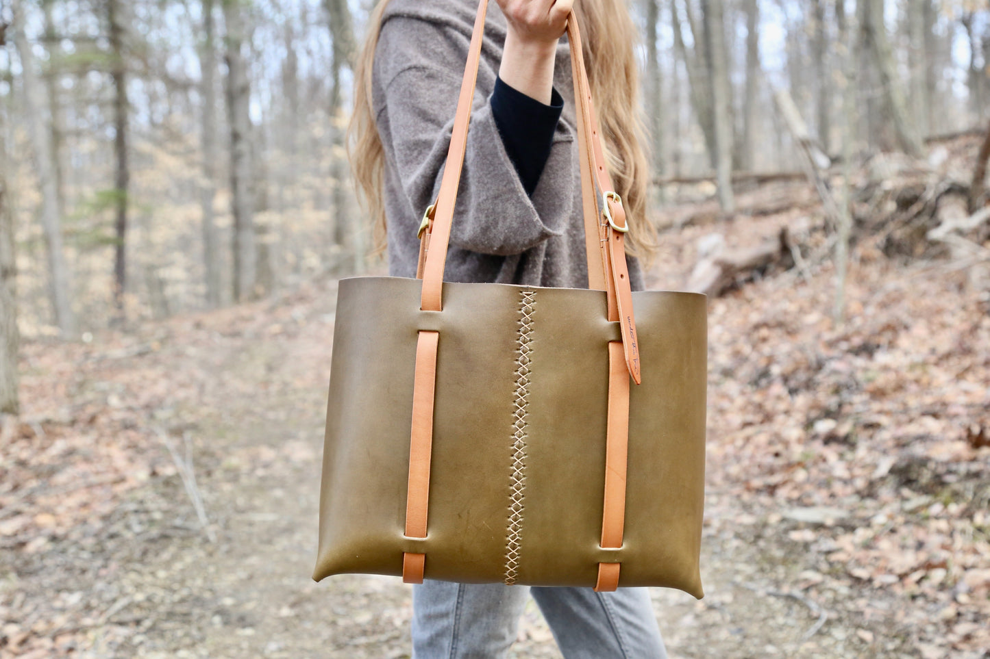 Crystal Van Gaasbeck from Under the Tree stands in the forest holding an olive green large tote bag with tan colored straps. She and her husband, Scott, make handmade leather bags and accessories in their hilltop studio in Willseyville, NY